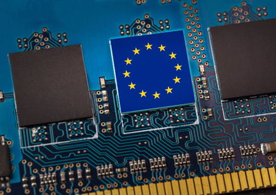 Close-up photograph of a computer circuit board with the European Union flag (blue background with circle of yellow stars) prominently displayed on a chip among other black components, representing EU technology regulation.